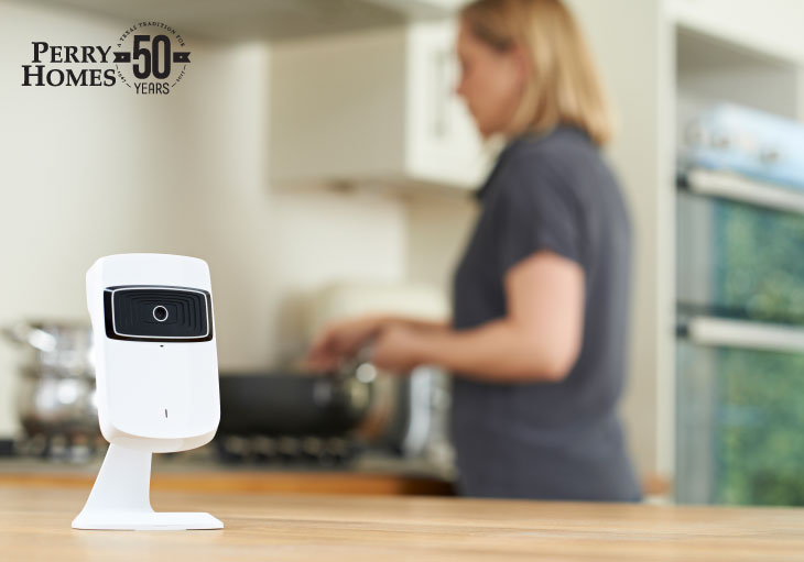 white and black smart security camera sits on wood kitchen countertop as woman in background cooks with pan on stove