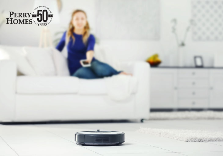 black circular robotic vacuum sits on white tile floor while woman sitting on couch in background points remote control at it
