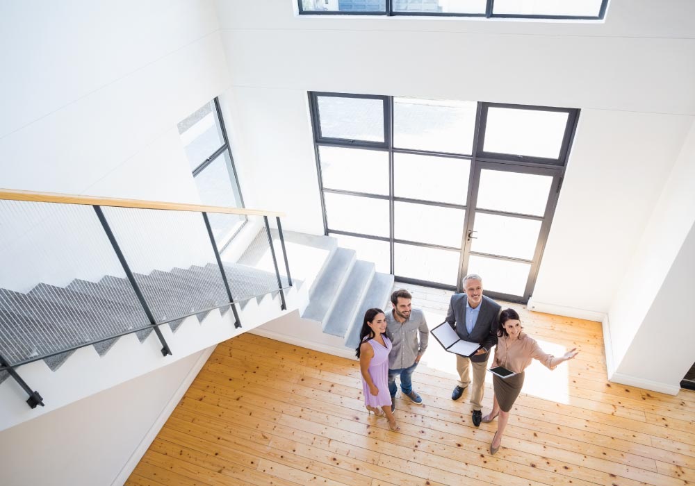 real estate agents showing young couple modern space with white walls, large windows, carpeted stairs and wooden flooring