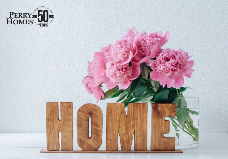 wood carving spelling out 'home' next to glass of pink peonies with subtle light grey and white background