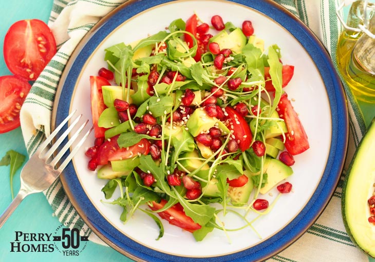 fork rests on white plate with blue border holds green salad with red tomatoes, pomegranite seeds and other green vegetables