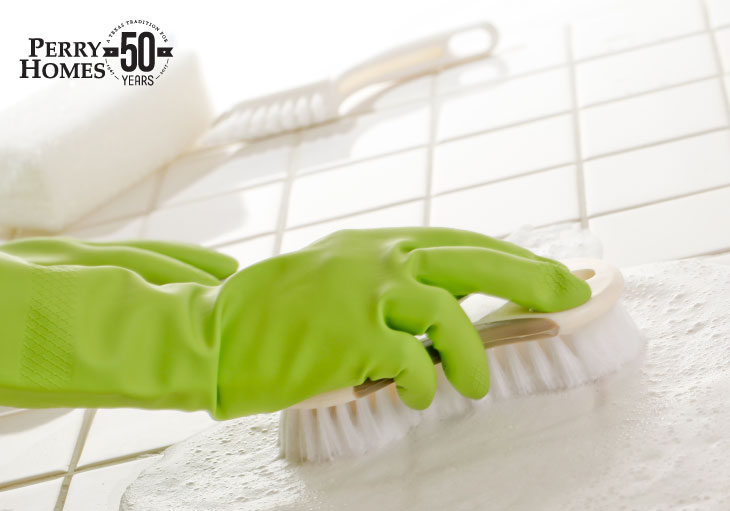 green gloved hand holding white scrub brush rubs soap on white tile  with other cleaning tools in background