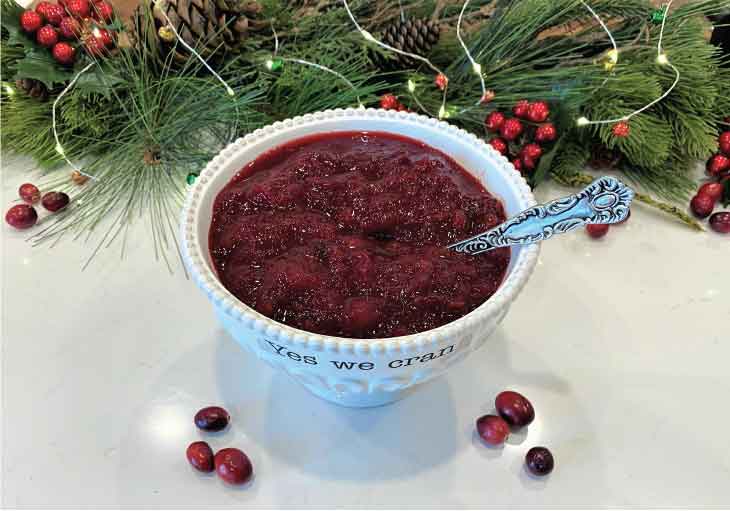 White ceramic bowl filled with cranberry sauce sits on white countertop decorated with holiday accessories.