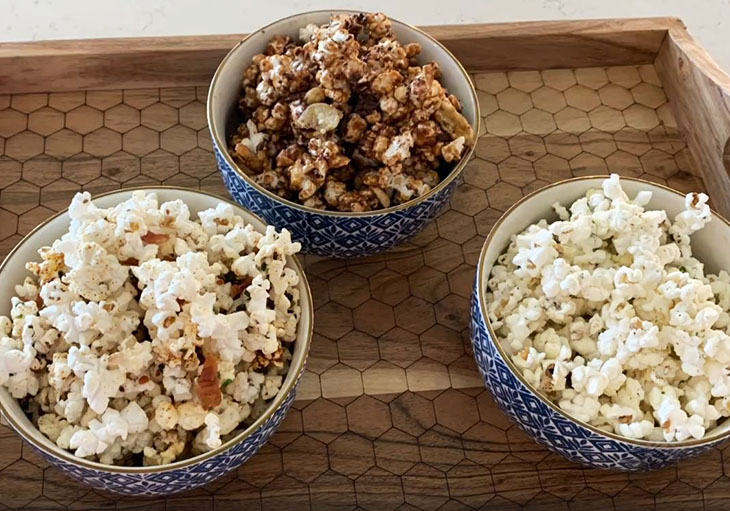 Close up photo of three blue ceramic bowls sitting on a wooden tray, that contain popcorn prepared with different seasonings.