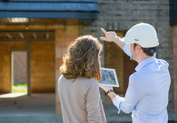 man wearing hard hat points to house under construction as he explains images on ipad to woman