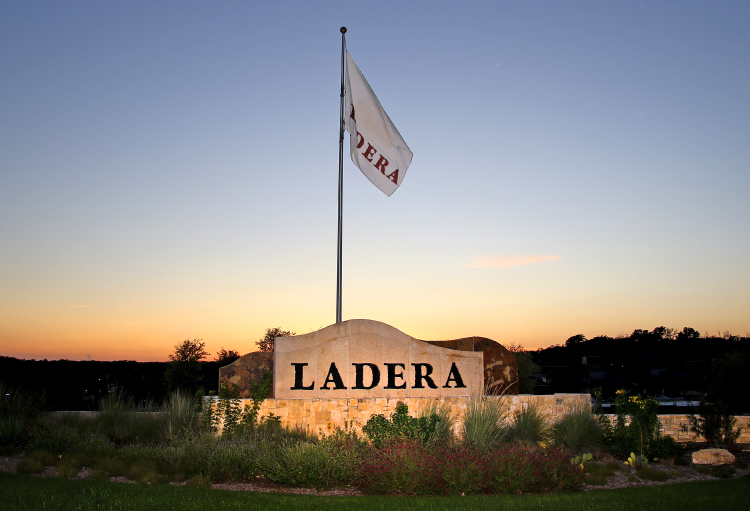 The sun sets on the Ladera communities sign and flag, which is surrounded by green bushes and trees.