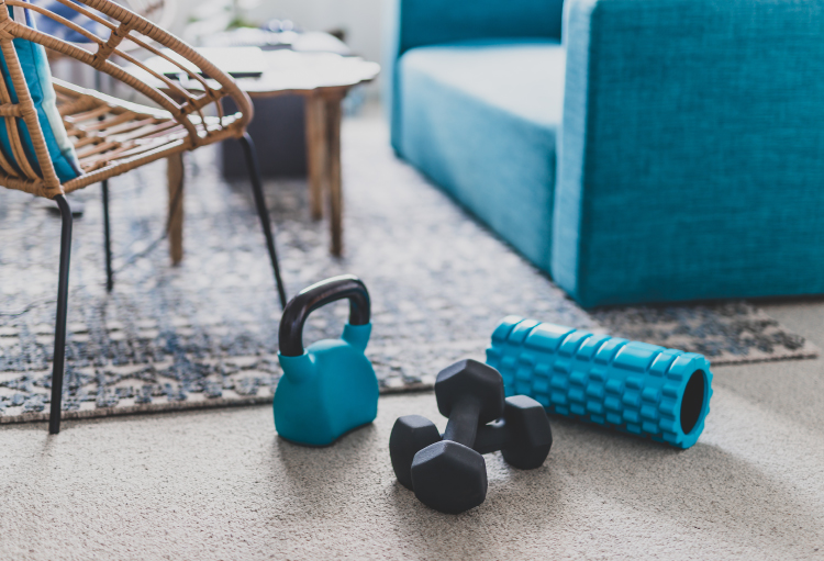 A kettlebell, dumbbell and back roller sit in a spacious, carpeted living room surrounded by furniture.