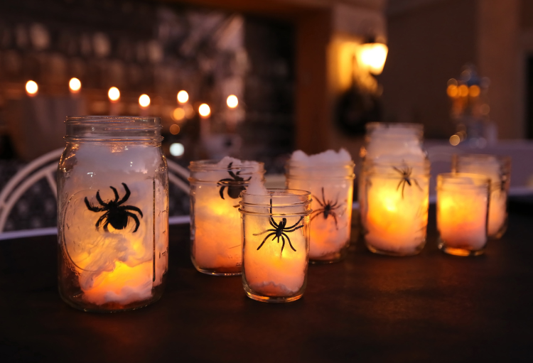 A series of mason jars sit on a table filled with cotton, lights and plastic spiders.