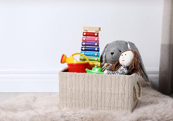 A small toy storage box made of a cream-colored knit material containing a colorful xylophone, grey rabbit, ragdoll and other miscellaneous toys sits atop a light tan fur rug in front of a white wall.