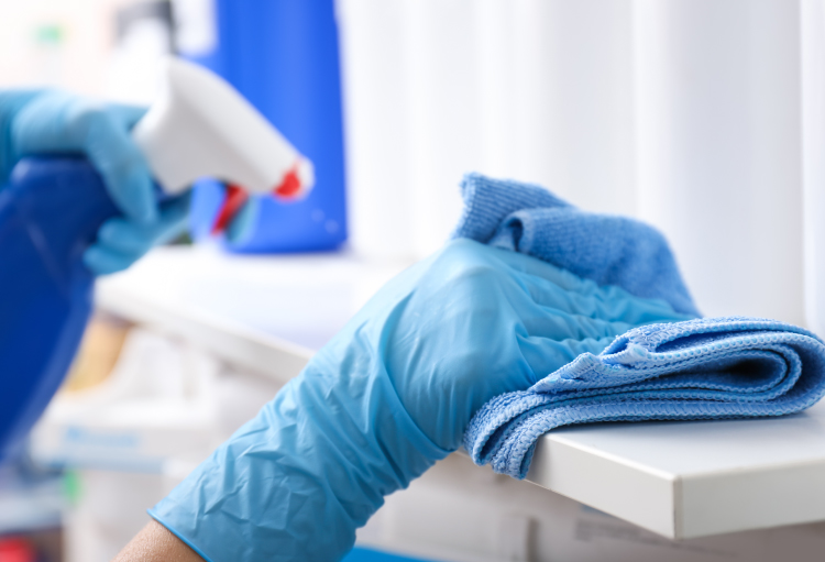 A person wearing blue latex gloves sprays cleaner on a floating shelf and wipes it with a blue terry cloth towel.