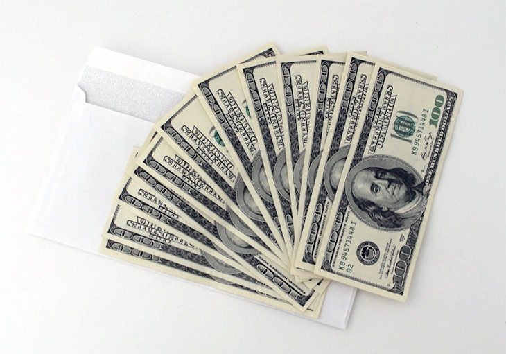 A stack of twelve $100 bills is fanned out over an open envelope on a white background.
