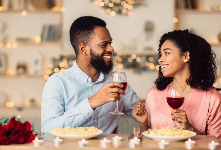 Young smiling couple raises wine glasses and eats homecooked meal during at-home Valentine’s Day date.